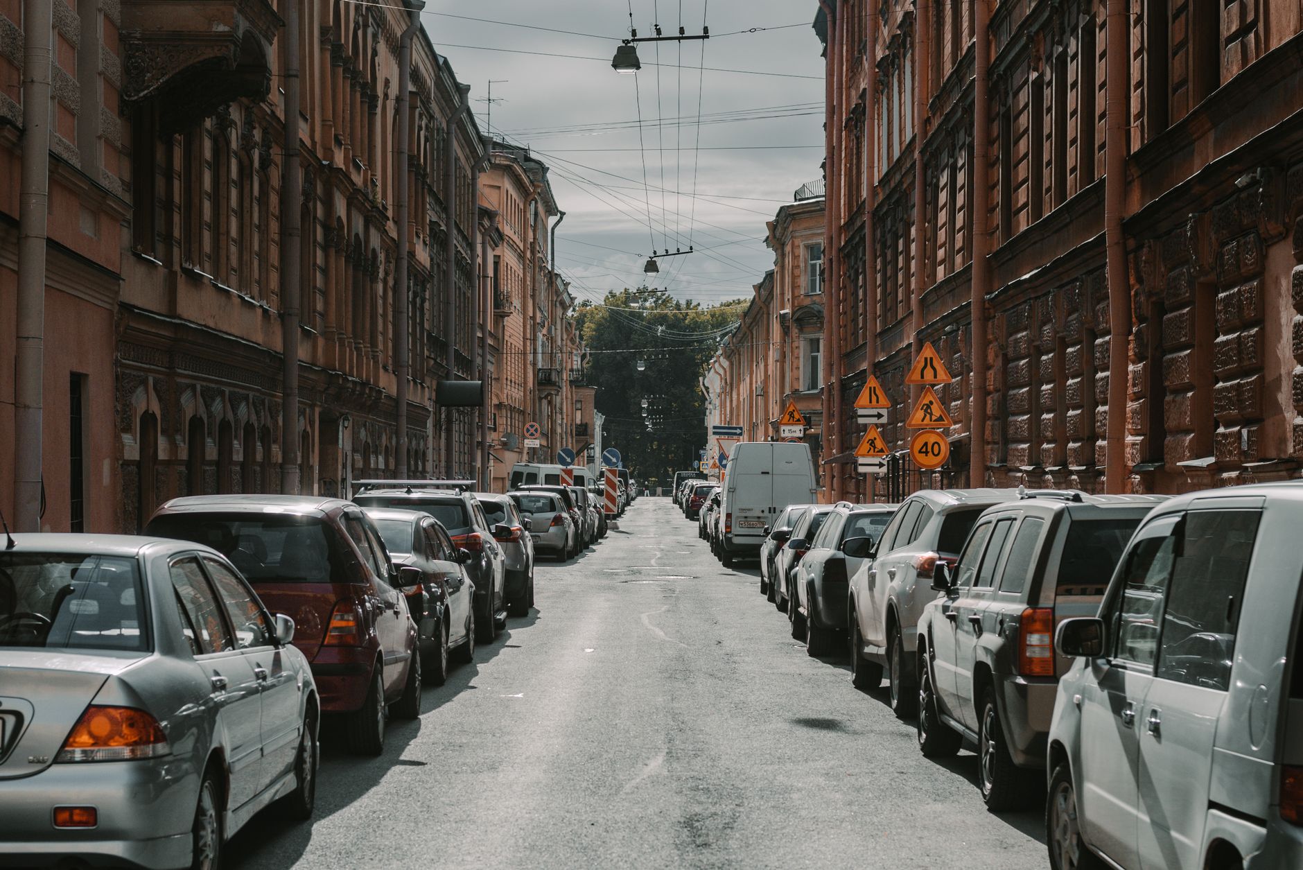 Long thin wires over narrow asphalt road surrounded by rows of cars under tall multi storey historical constructions in dayti