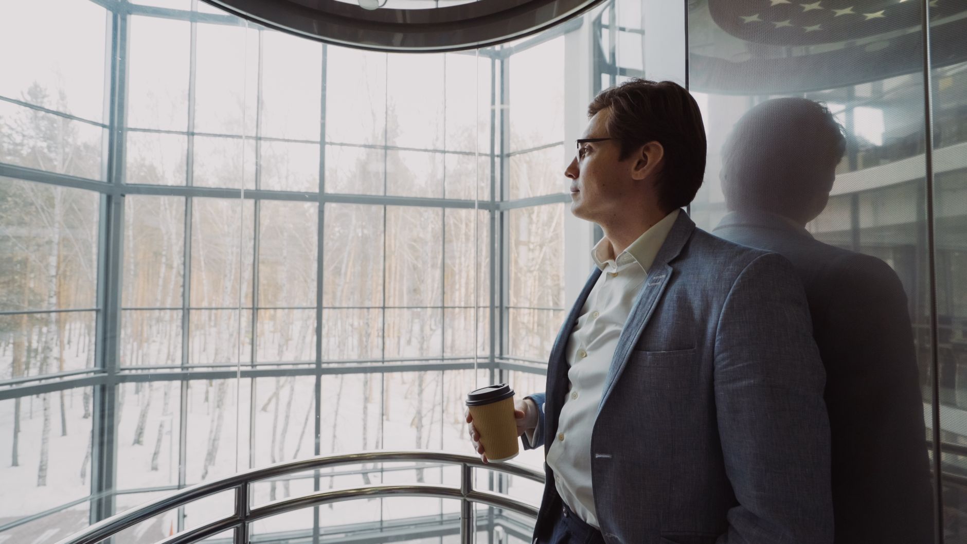 Businessman in blue suit with coffee cup looking out a large window, reflecting on a winter landscape. — Ever-Growing List of