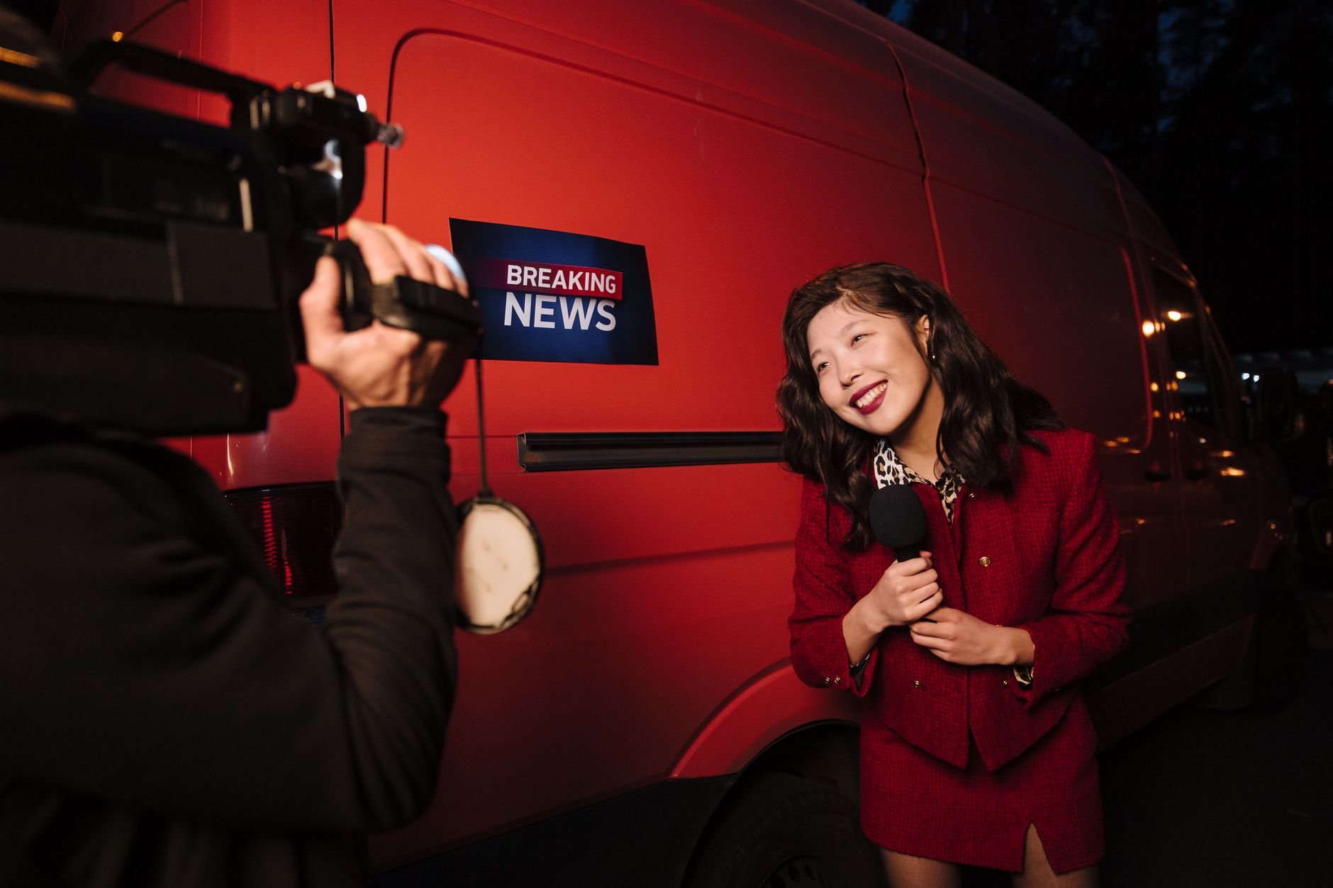 Asian female reporter in a red blazer reporting live on the news outdoors in the evening. — Hello world!
