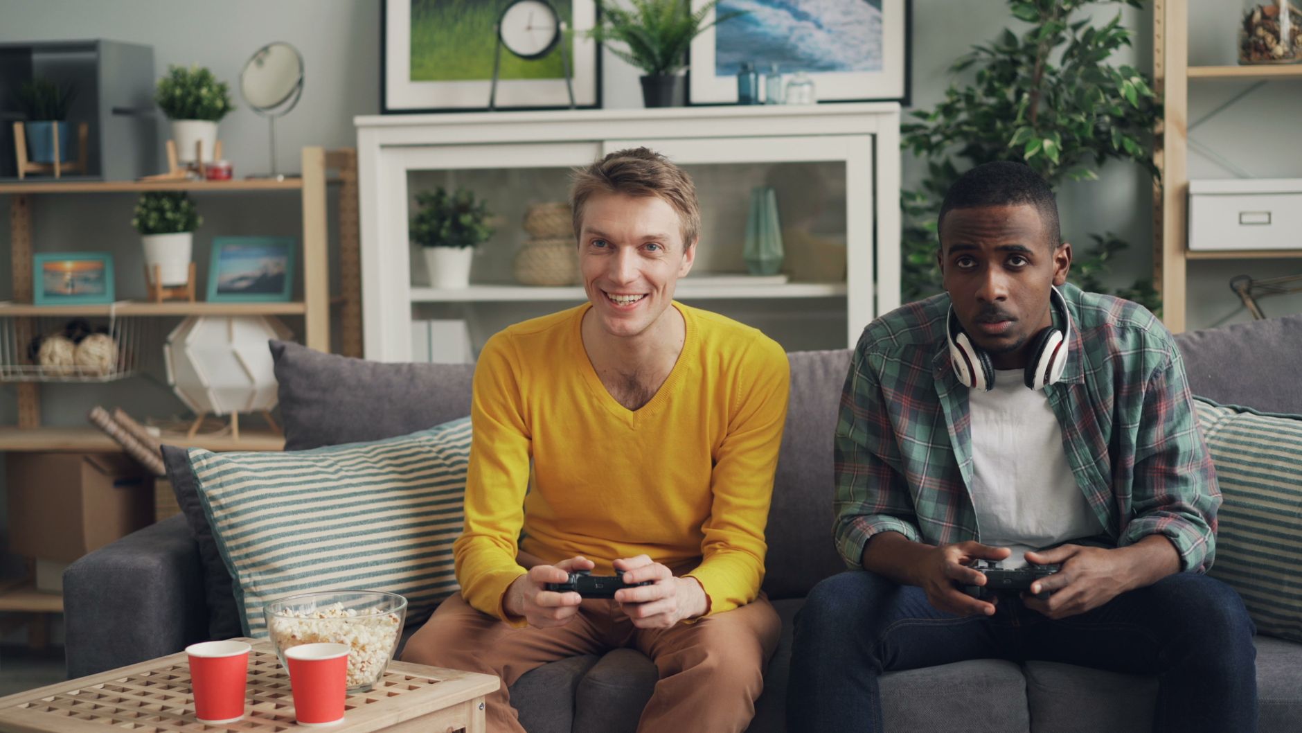 Two friends playing video games together on a sofa, one holding a controller and the other wearing headphones. — Pick The Bes