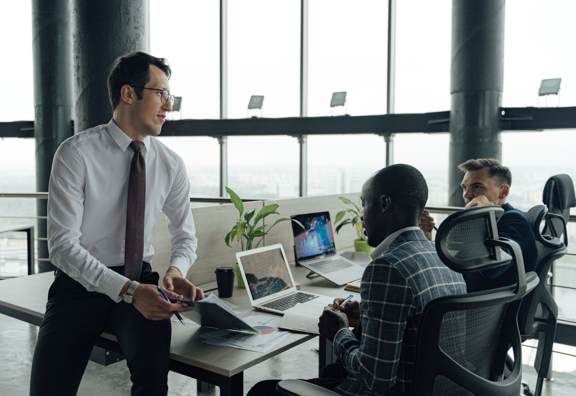Three men engaged in a business meeting in a modern office setting with laptops and documents. — Understanding the Uses and E